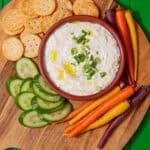 knorr spinach dip topped with chopped onions and a drizzle of olive oil in a red bowl on a cutting board and surrounded by carrots, cucumbers, and crackers