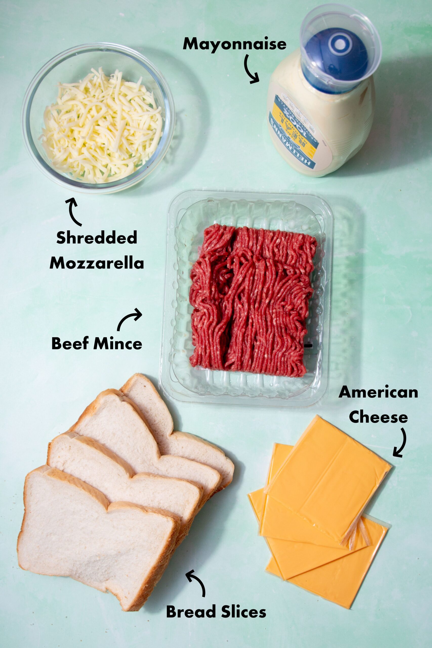 Ingredients to make the grilled cheese burger in a skillet laid out on a pale blue background and labeled with arrows.
