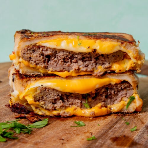 grilled cheese burger that has been cut in half and then the halves stacked on top of each other, laying on a wooden cutting board near green leaves.