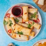 plate of air fryer spring rolls and a cup of soy sauce sprinkled with red chilli slices and cilantro on a blue background