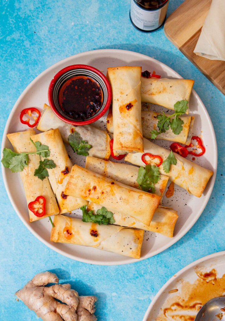 plate of air fryer spring rolls and a cup of soy sauce sprinkled with red chilli slices and cilantro on a blue background