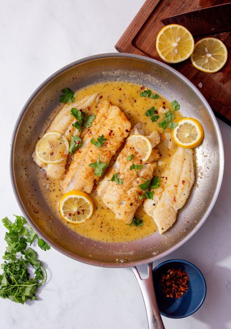 Image of baked flounder recipe in a skillet with white wine sauce and lemon. Topped with fresh parsley