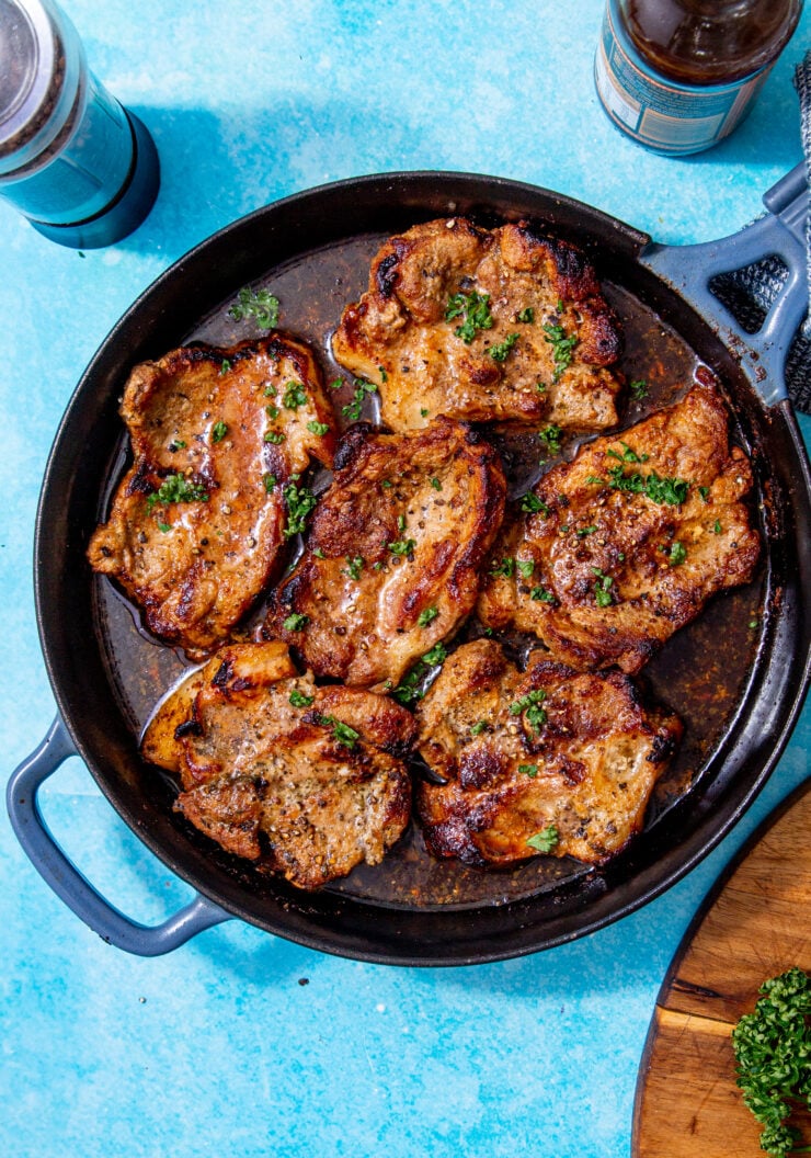 juicy, fully baked pork chops in a skillet that were baked in the oven and topped with fresh parsley all on a blue background