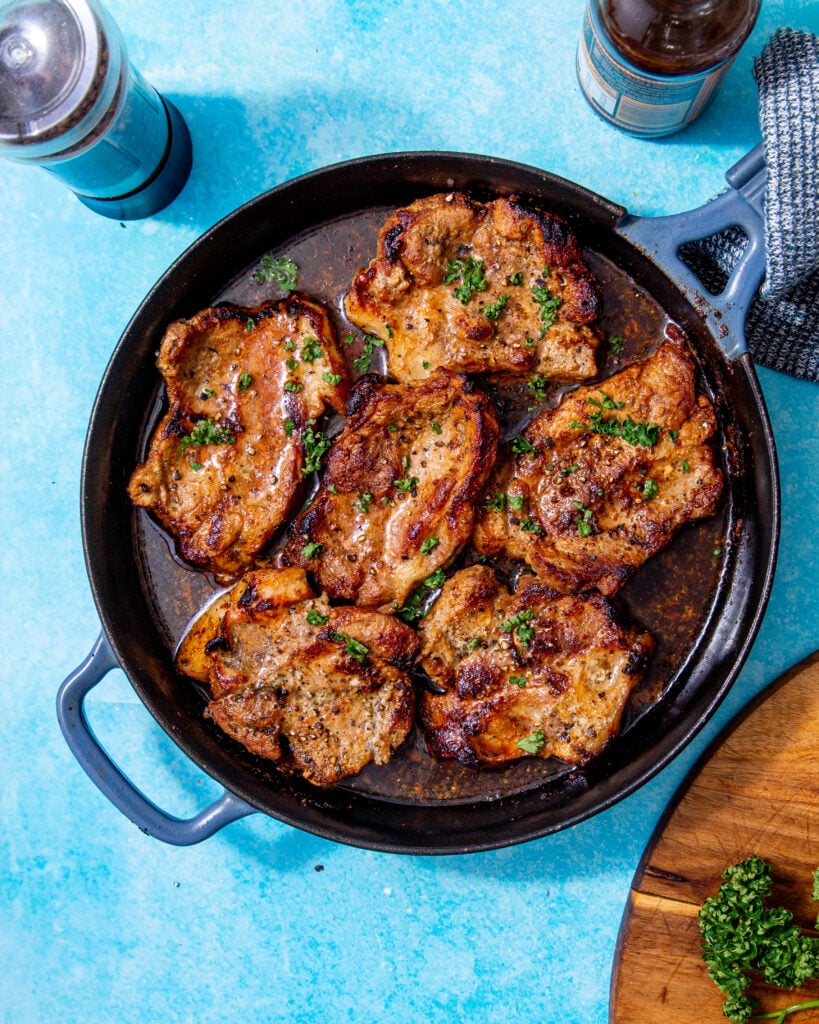 juicy, fully baked pork chops in a skillet that were baked in the oven and topped with fresh parsley all on a blue background