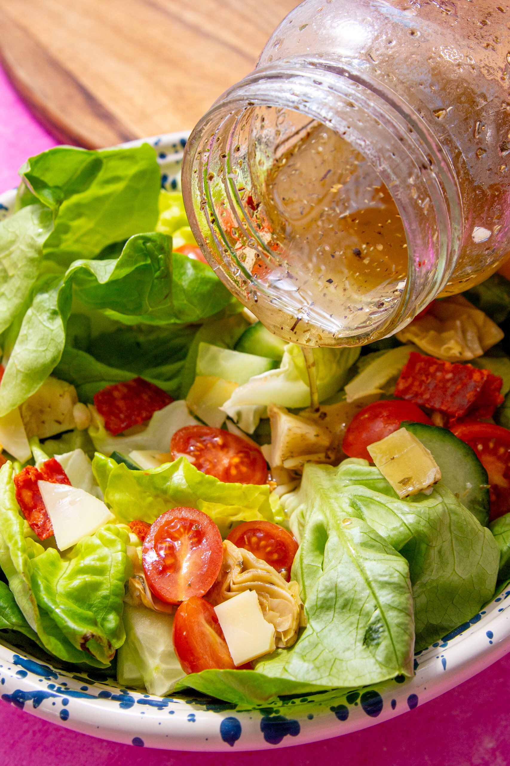 close up of prepared homemade submarine dressing being poured over a salad with tomatoes, Italian meats, artichokes, and cucumbers form a mason jar, all on a pink background