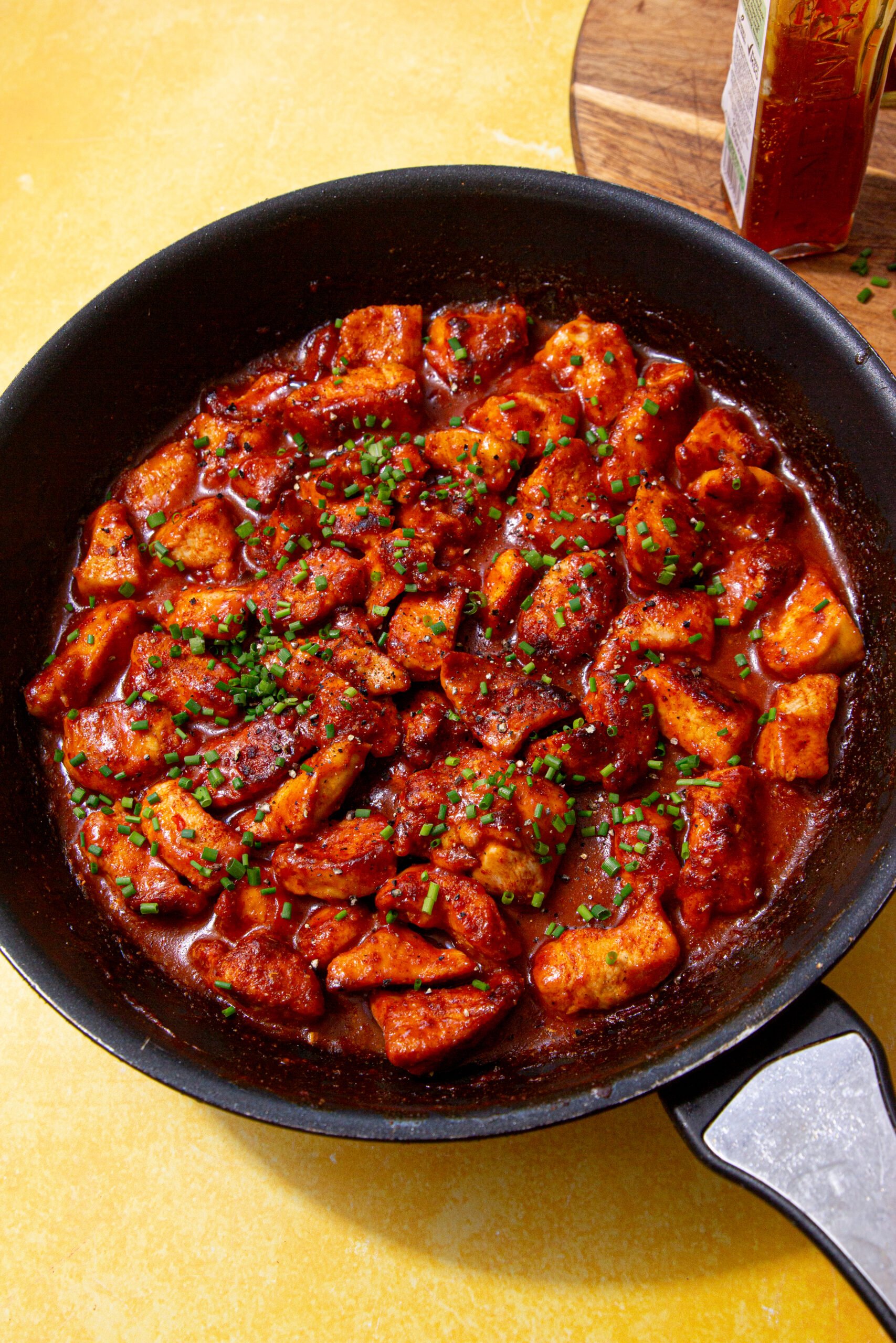 sweet chilli chicken in a black skillet on a yellow background next to a cutting board with a bottle of sweet chilli sauce on it.