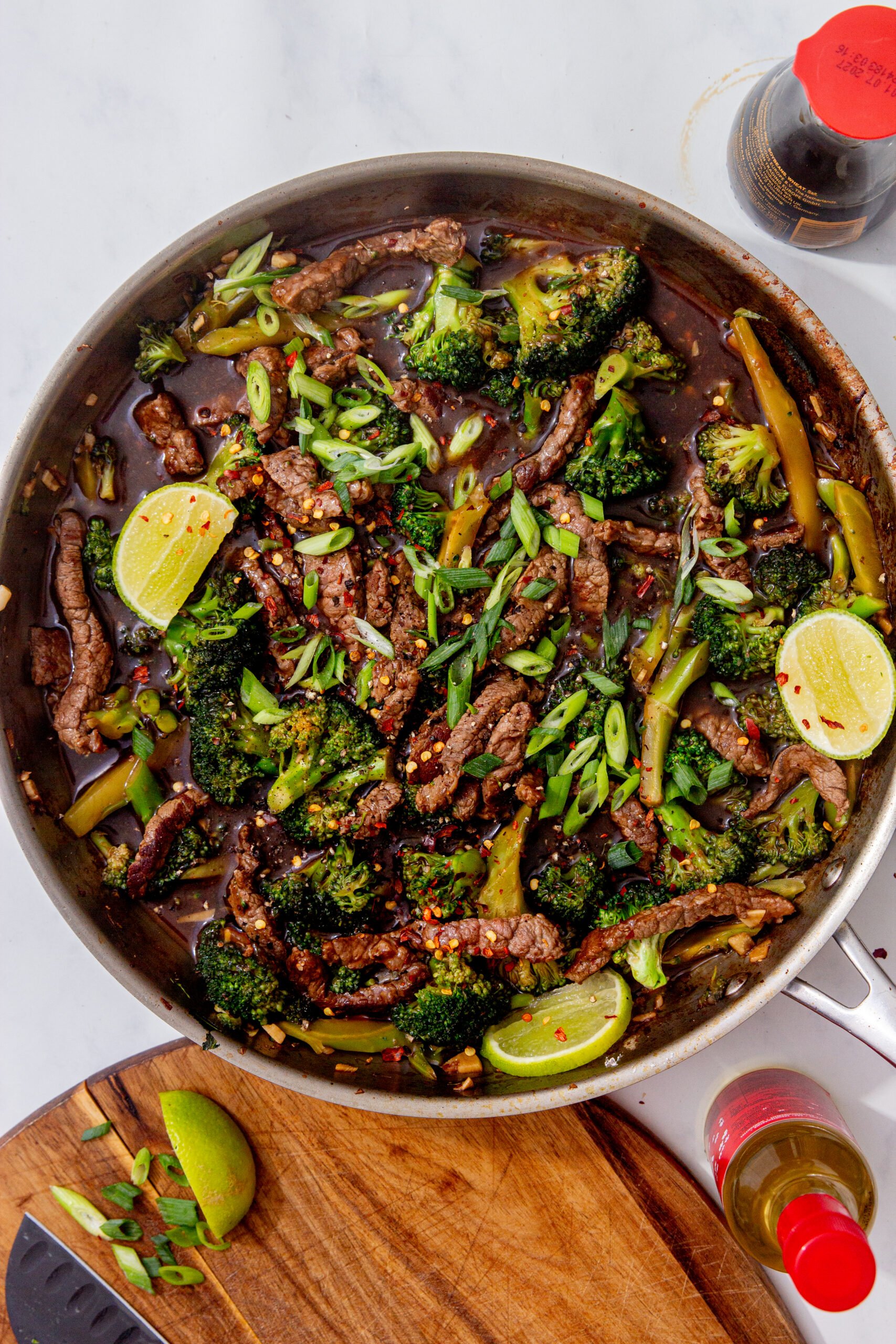 beef and broccoli recipe in a skillet on a white background and garnished with lime wedges