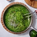 A pair of tongs in prepared green spinach pasta sauce served with spaghetti and topped with parmesan cheese and chili flakes in a stainless steel pan
