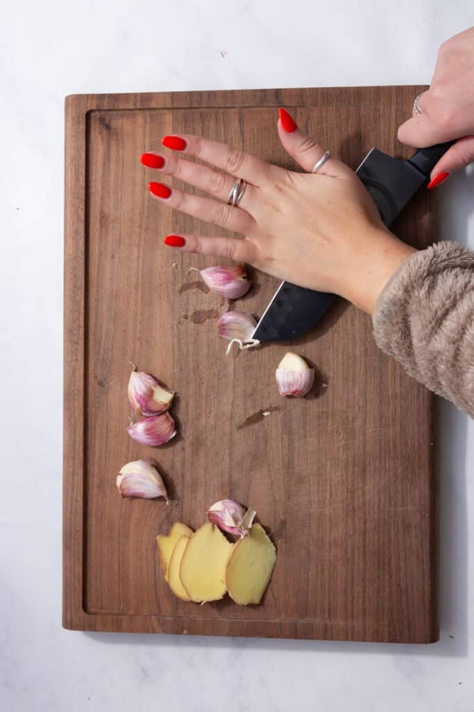 Garlic cloves being smashed next to sliced ginger on a cutting board.  and the second showing a whole chicken in a stock pot with other ingredients.
