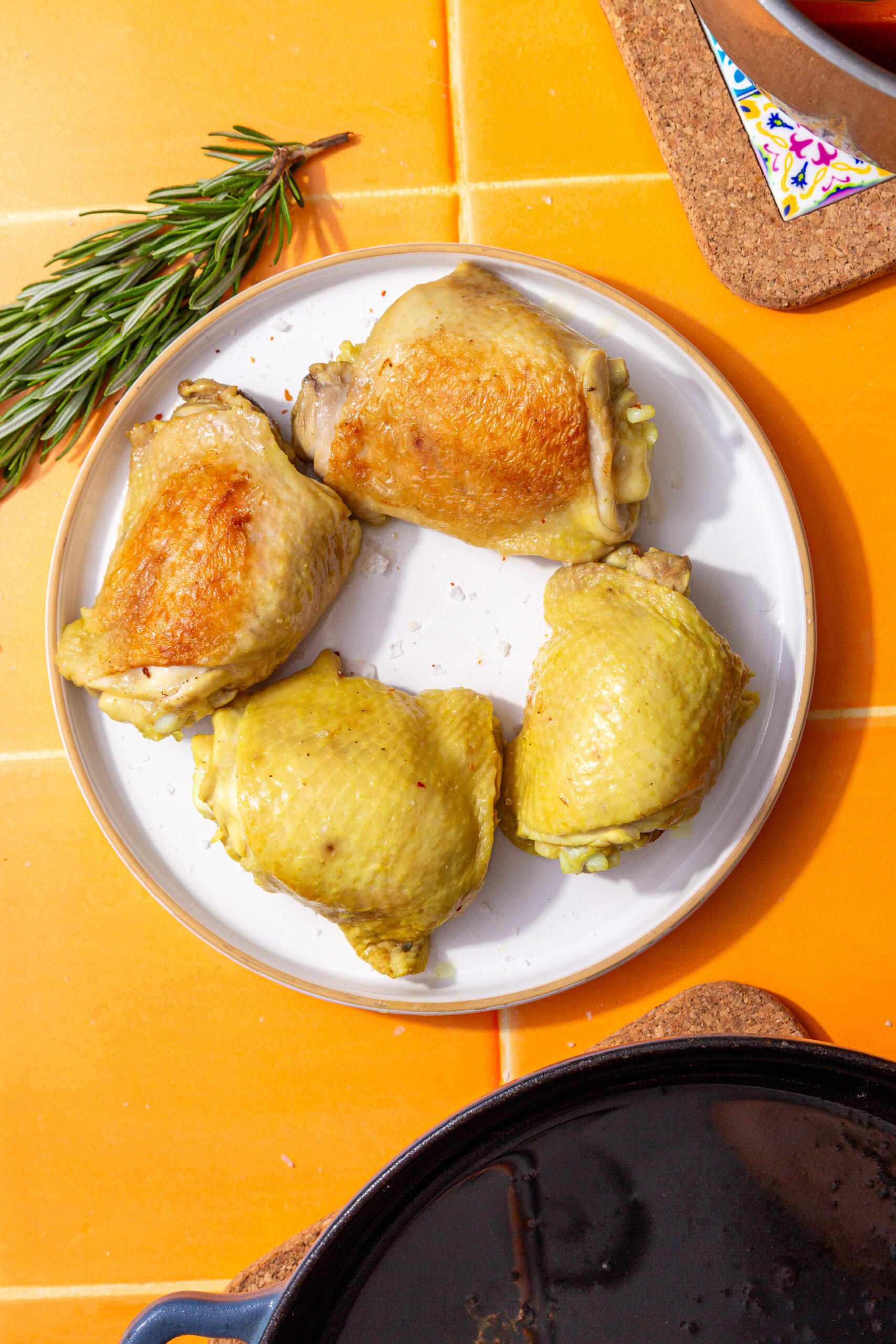 Fully boiled chicken thighs on a white plate set on orange tile next to a sprig of rosemary.
