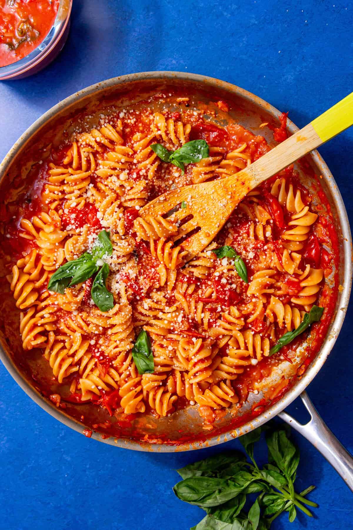Spiral noodles covered with red sauce and sprinkled with white parmesan cheese all in a silver pan with a tan wooden spoon in the pan.  Garnished with green basil leaves.