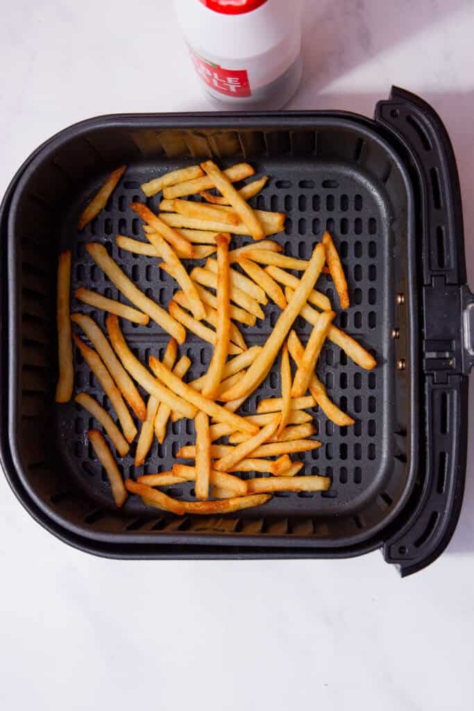 Golden French fries are spread out in the basket of a black air fryer on a white countertop, with a partially visible salt shaker at the top of the image.