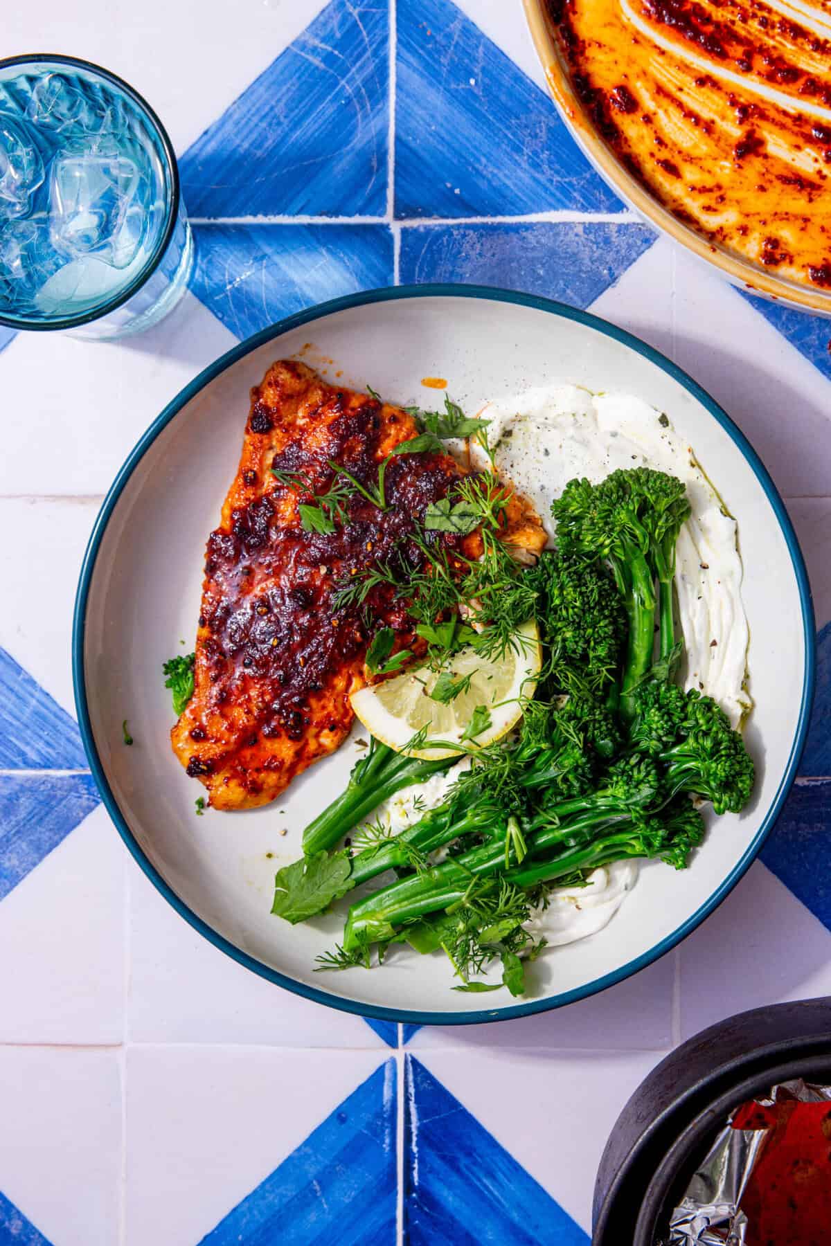 Cod fillet with a red seasoning mixture plated on a white plate with a blue rim next to tenderstem broccoli and garnished with fresh herbs and a lemon wedge.