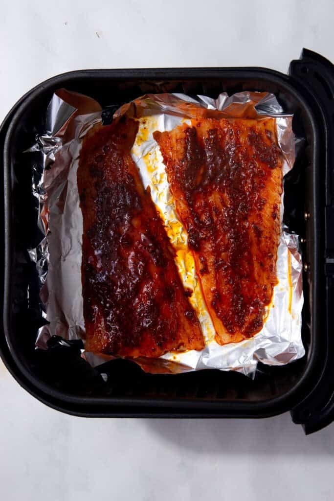 An air fryer basket that has been lined with foil and has two fish fillets on top that have a reddish brown seasoning on them.