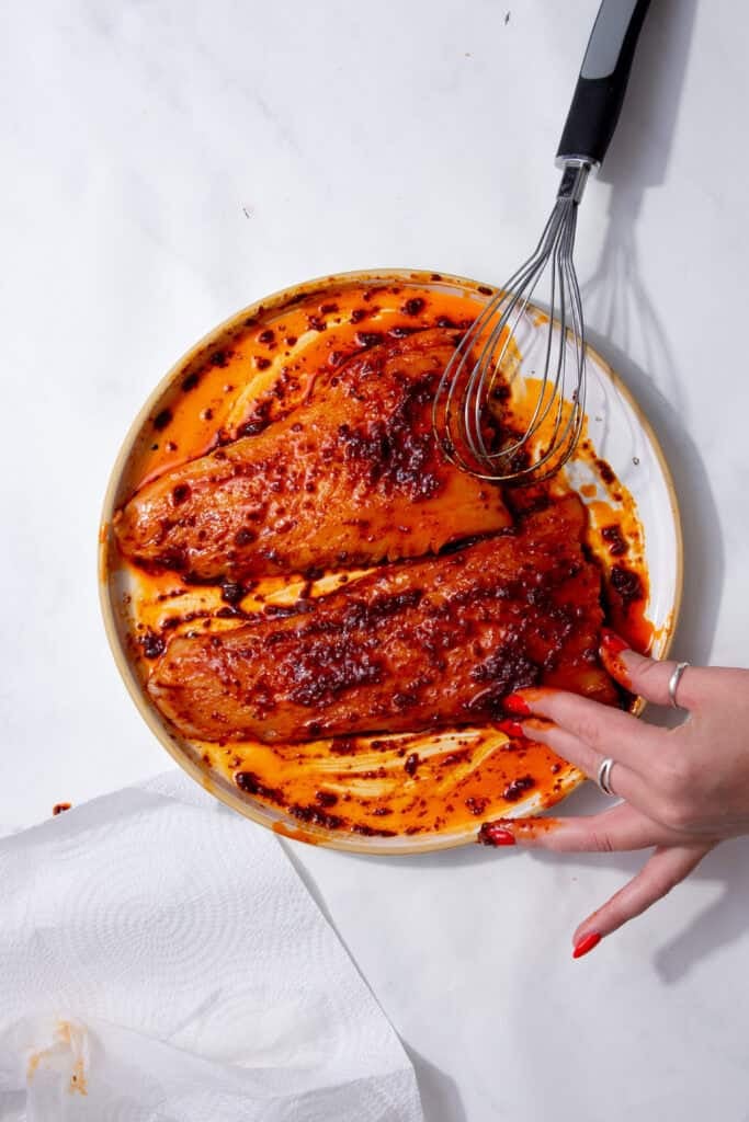 A hand with red nails holding the side of a cod fillet that has reddish brown mixture all over it and a whisk to the side.