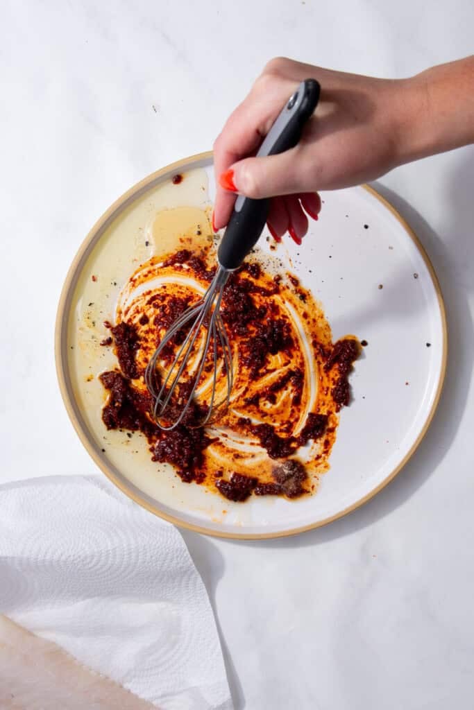A hand using a whisk to mix together a reddish brown paste on a white plate.