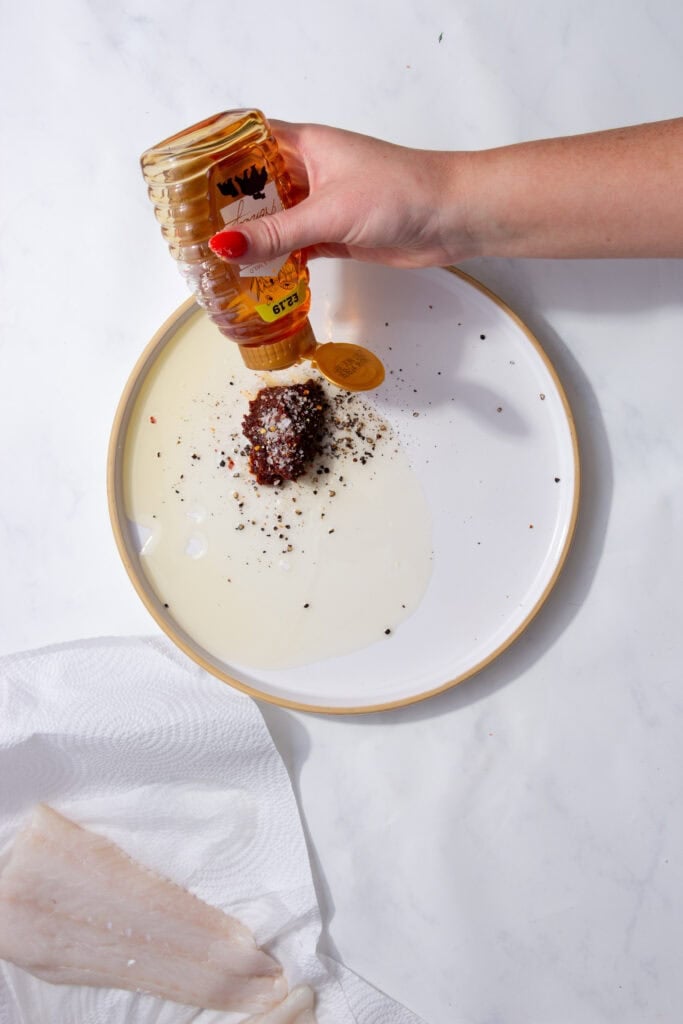 A hand squeezing honey onto a plate with a red ball of seasoning.