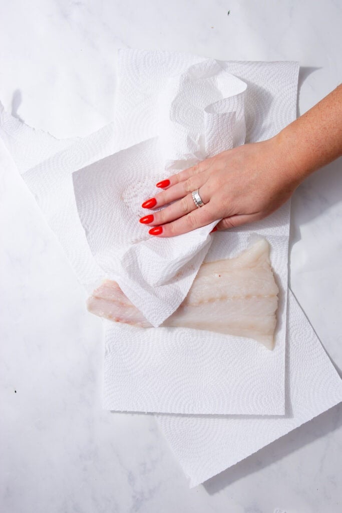 A hand with red nail polish and a silver ring patting cod fillets dry with a paper towel.