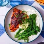 Well seasoned cod fillet on a white plate with tenderstem broccoli and a lemon wedge all on a blue and white tile background.