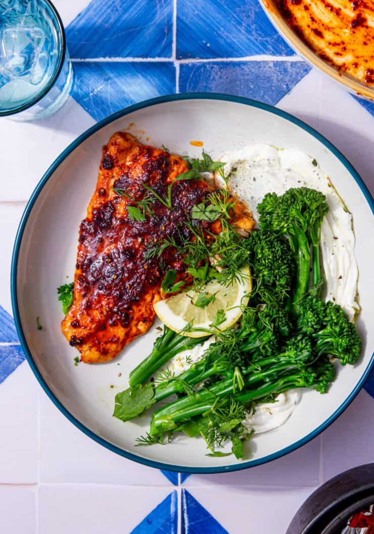 Well seasoned cod fillet on a white plate with tenderstem broccoli and a lemon wedge all on a blue and white tile background.