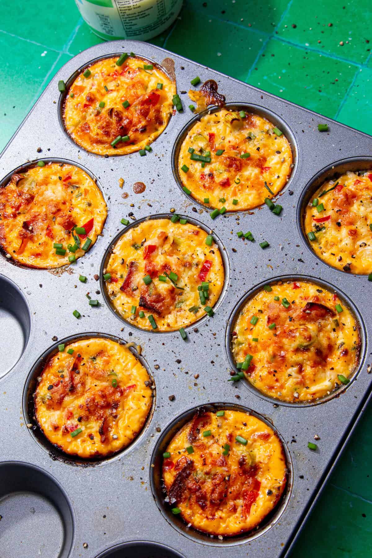 Image of baked yellow egg bites with a browned top in a grey muffin tin and sprinkled with chopped chives all on a green tile background.