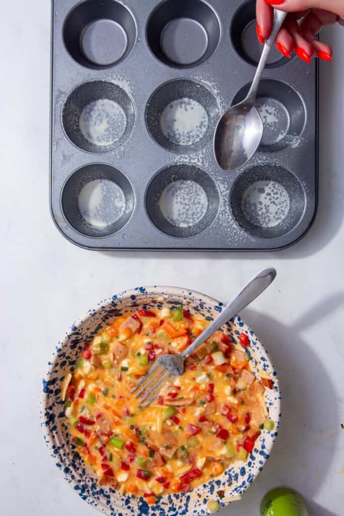 A colorful mixture of eggs and vegetables in a blue and white speckled bowl next to a grey muffin tin.