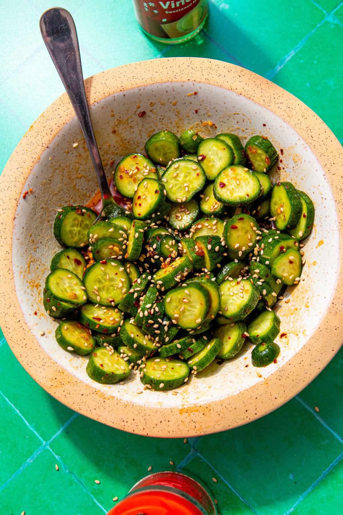 Sliced cucumbers topped with red chili sauce and sesame seeds with a spoon sticking out and all in a white and tan bowl on a green background.