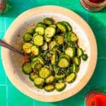 bowl of sliced cucumbers with seasoning sprinkled on top and a metal spoon sticking out all in a white and tan bowl on a green background.