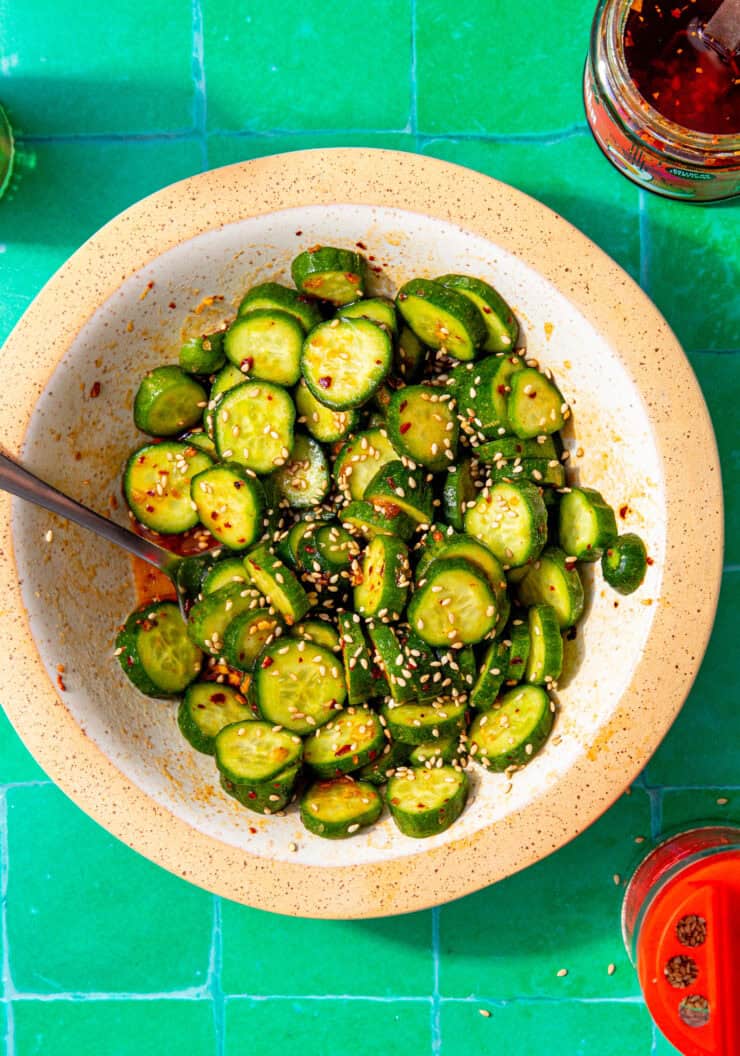 bowl of sliced cucumbers with seasoning sprinkled on top and a metal spoon sticking out all in a white and tan bowl on a green background.