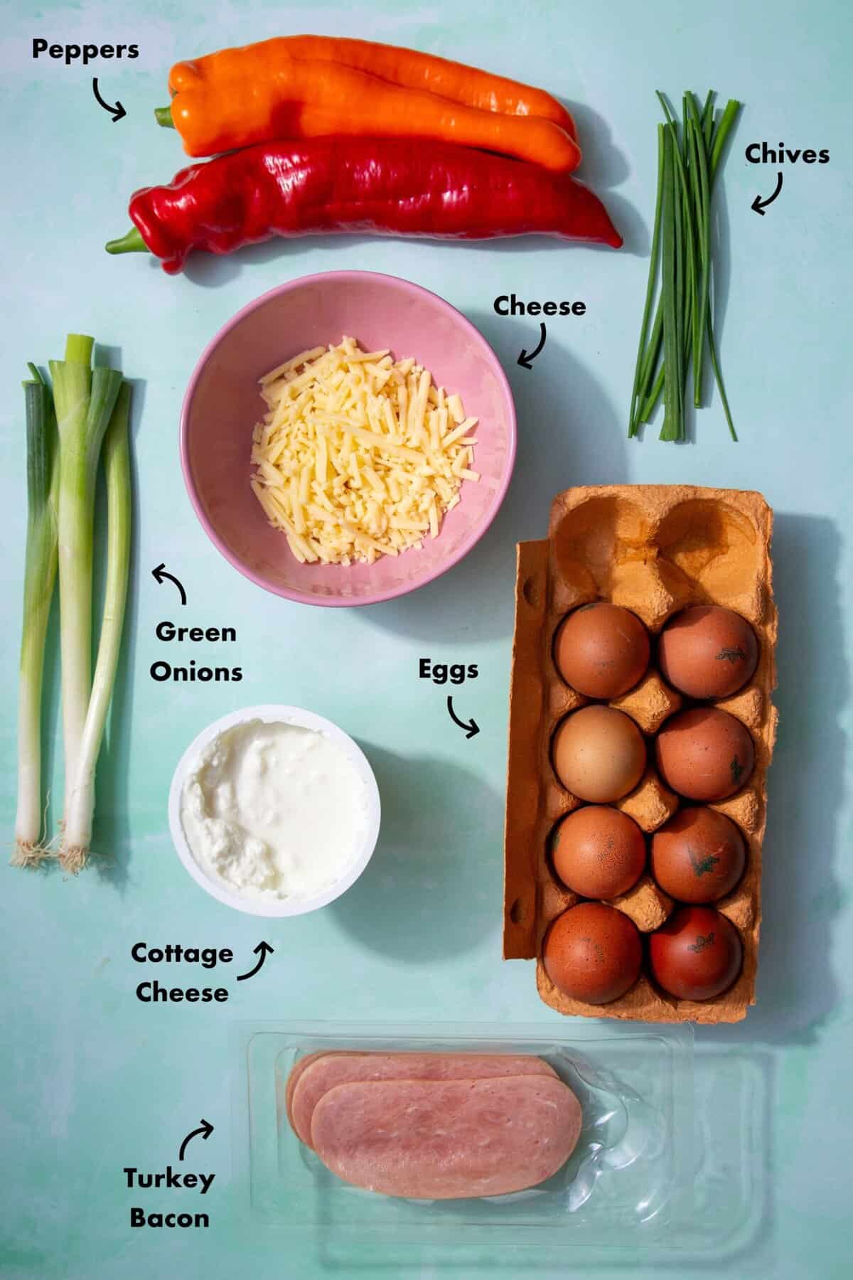 Ingredients to make the cottage cheese egg bites in the oven laid out on a pale blue background and labeled with arrows.
