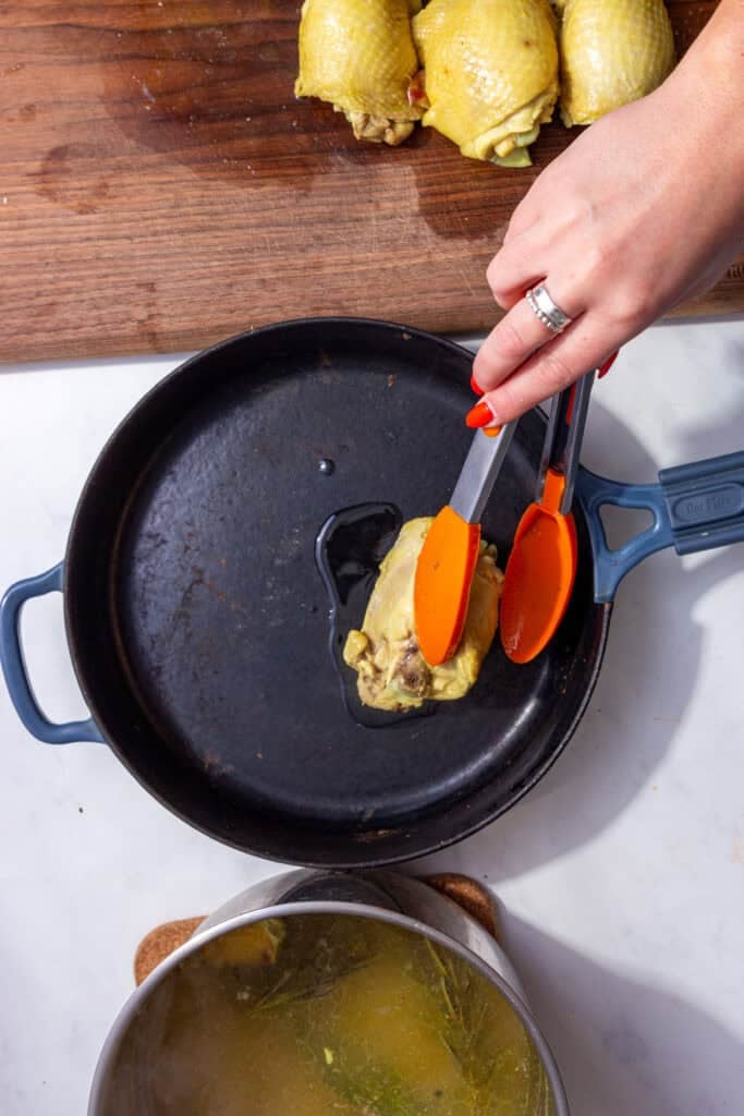 A boiled chicken thigh being added to a cast iron pan with oil by a pair of tongs being held by a hand.
