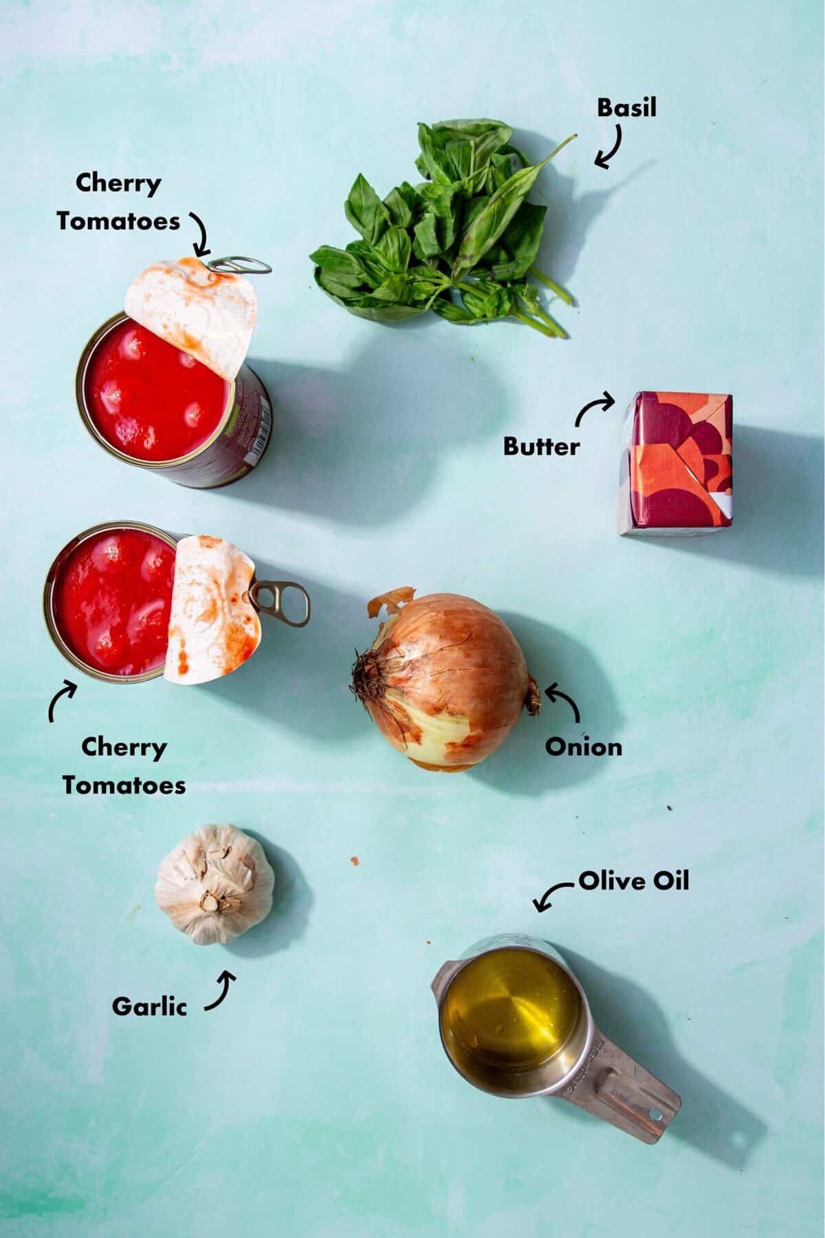Ingredients to make the tomato basil sauce in a skillet laid out on a pale blue background and labeled with arrows.
