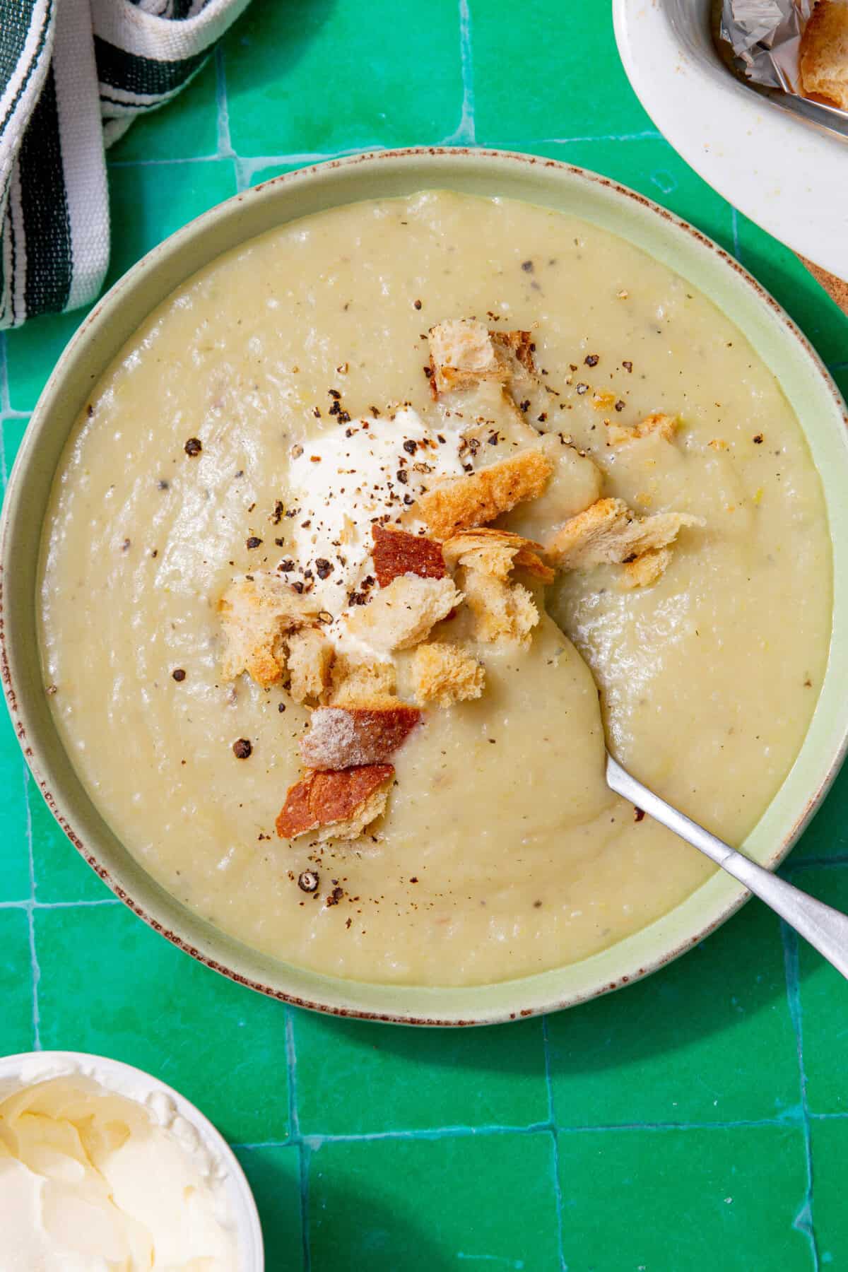 An image of tan soup in a bowl topped with white cream and croutons all on a green tile background.