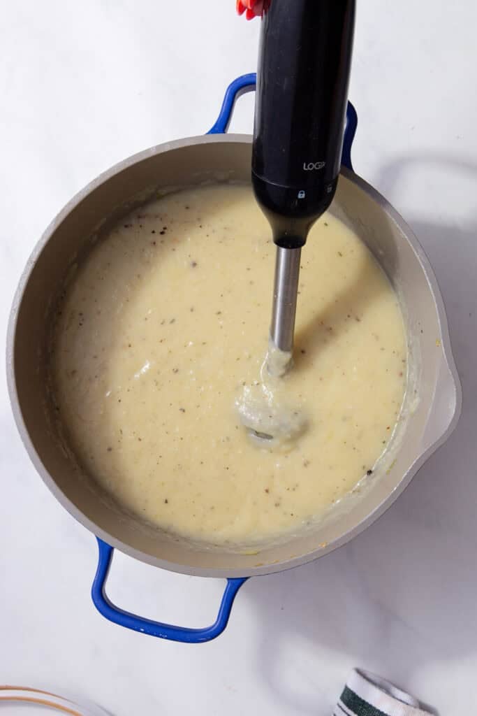 An immersion blender in a pan of creamy light yellow substance.