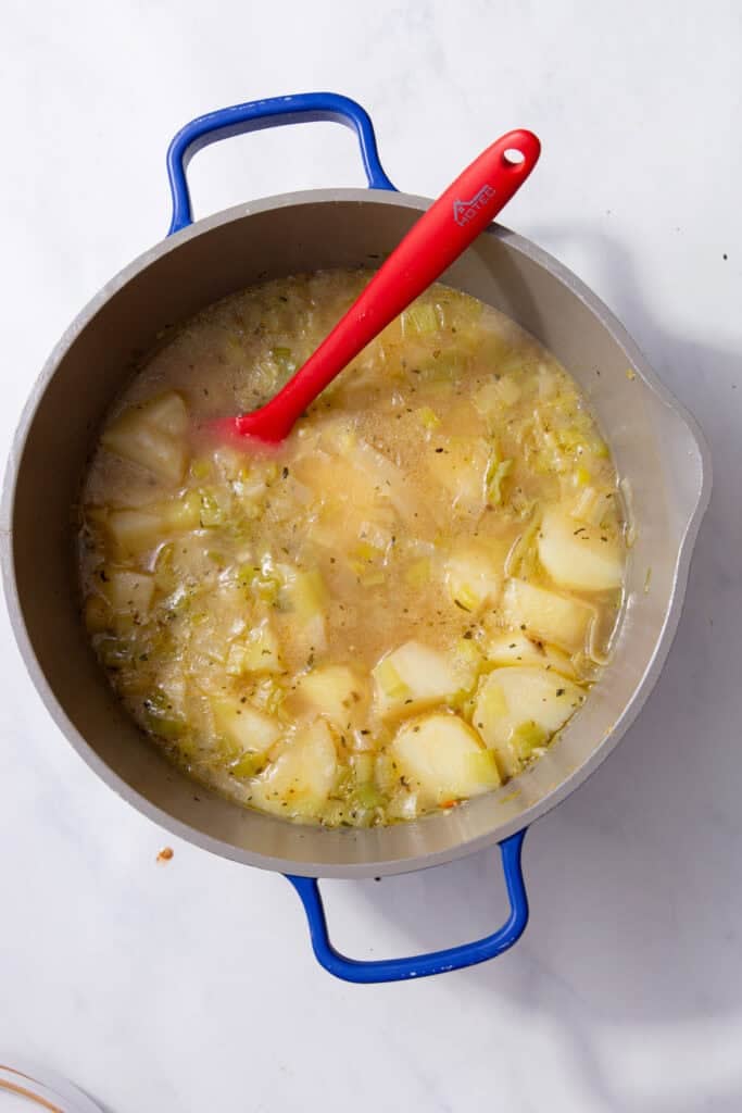 A red spatula in the silver pan with blue handles stirring the yellow liquid.