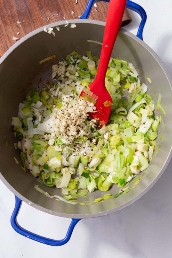 A red spatula stirring the green leeks and pepper in a silver pan with blue handles.