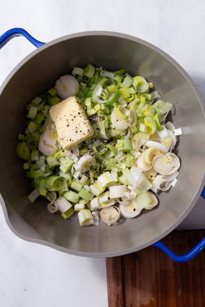 Green leeks, butter and pepper in a silver pan with blue handles.