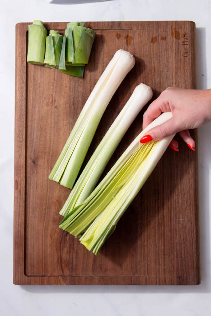 A hand with red nail polish holding the bottom part of a leek on a cutting board.