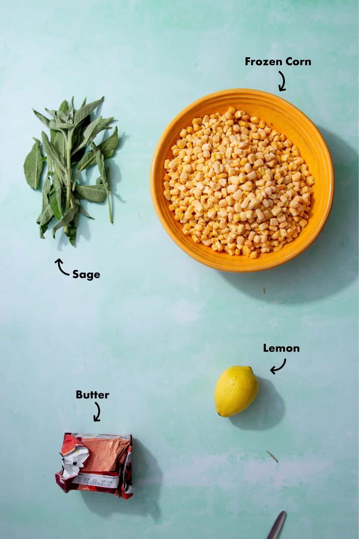 A bunch of green sage leaves, a yellow bowl filled with yellow corn kernels, a yellow lemon, and a red package of butter labeled with words and arrows on a light greenish blue background.