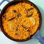 Image of roasted corn in a skillet with roasted sage leaves and lemon wedges on a light green background.