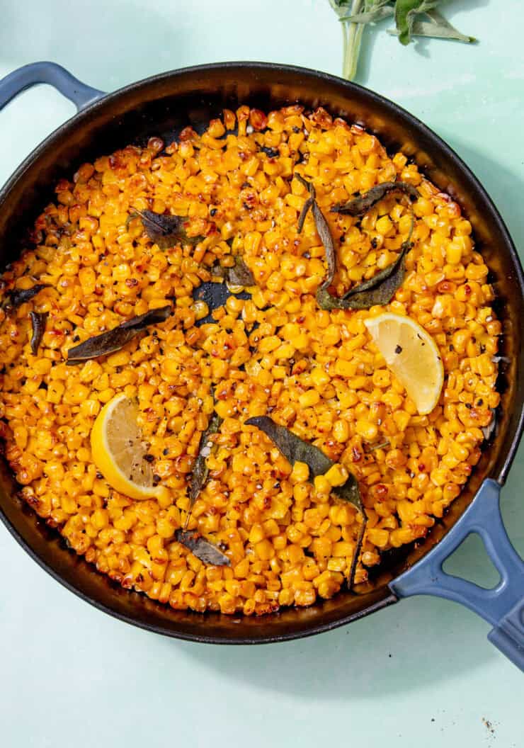 Image of roasted corn in a skillet with roasted sage leaves and lemon wedges on a light green background.