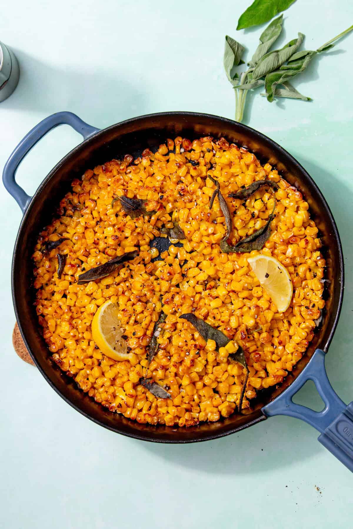 Roasted corn in a skillet with lemon wedges and cooked sage leaves.