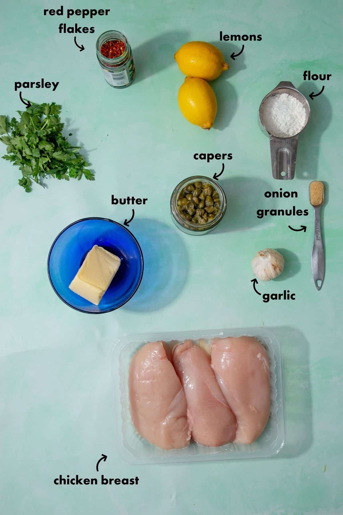 Ingredients laid out for easy chicken piccata, including chicken breast, lemons, garlic, capers, butter, flour, parsley and red pepper flakes.