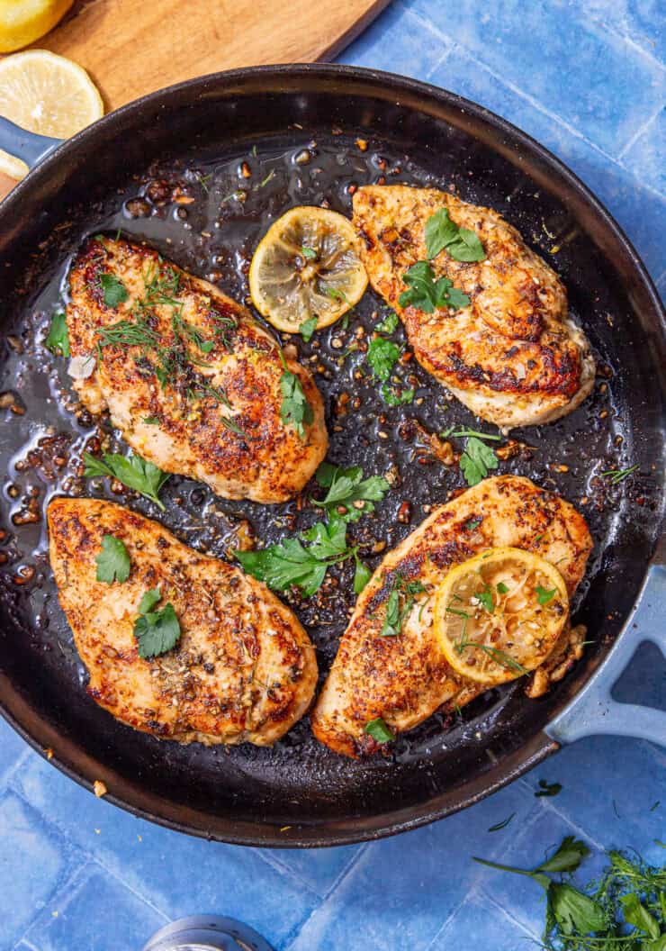 Tender cast iron chicken breasts seared to golden perfection in a skillet, garnished with fresh parsley, herbs, and lemon slices on a blue tile background.