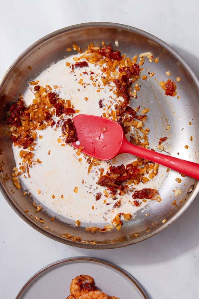 Cream being stirred into the onion and tomato base with a red spatula.