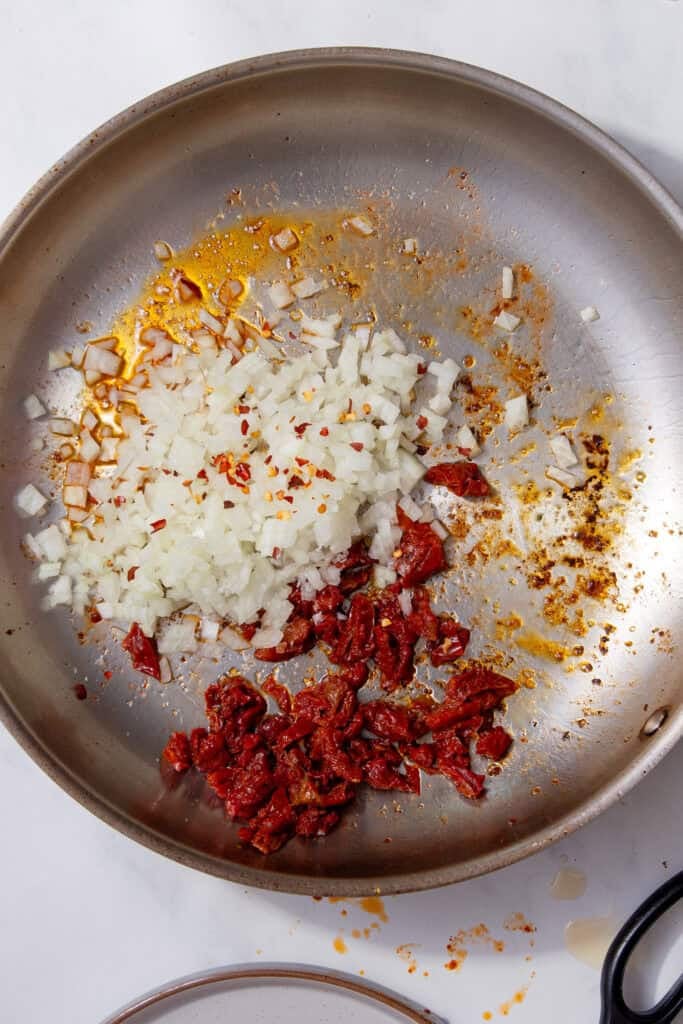 A pan with diced onion and chopped sun-dried tomatoes sizzling in oil.