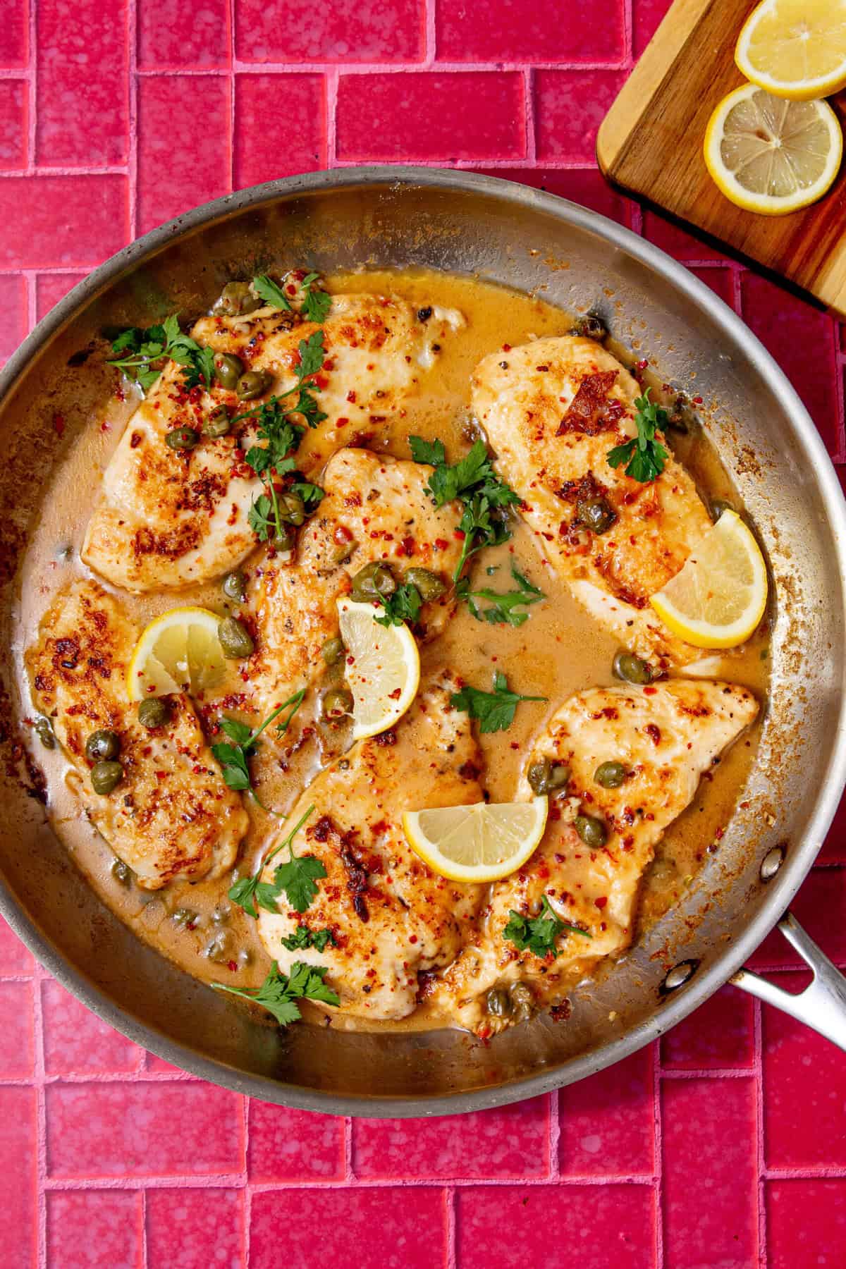 A pan of easy chicken piccata with lemon slices, capers and parsley on a red tiled background.