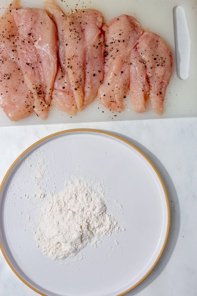 Seasoned raw chicken breasts on a board next to flour on a plate.