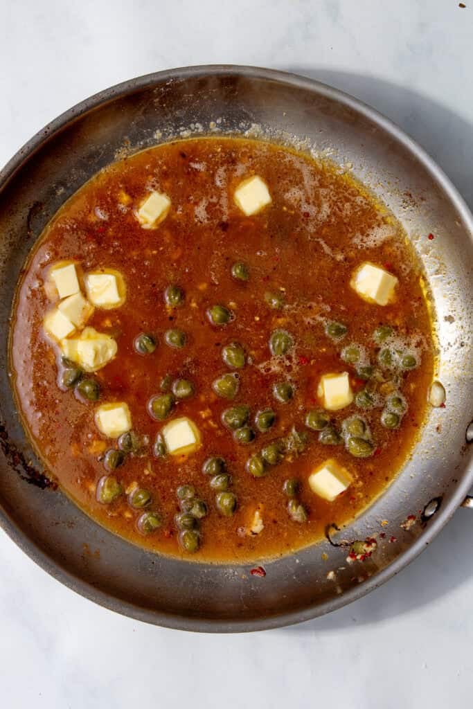 Chicken broth and capers simmering in the pan with cubes of butter melting on top.