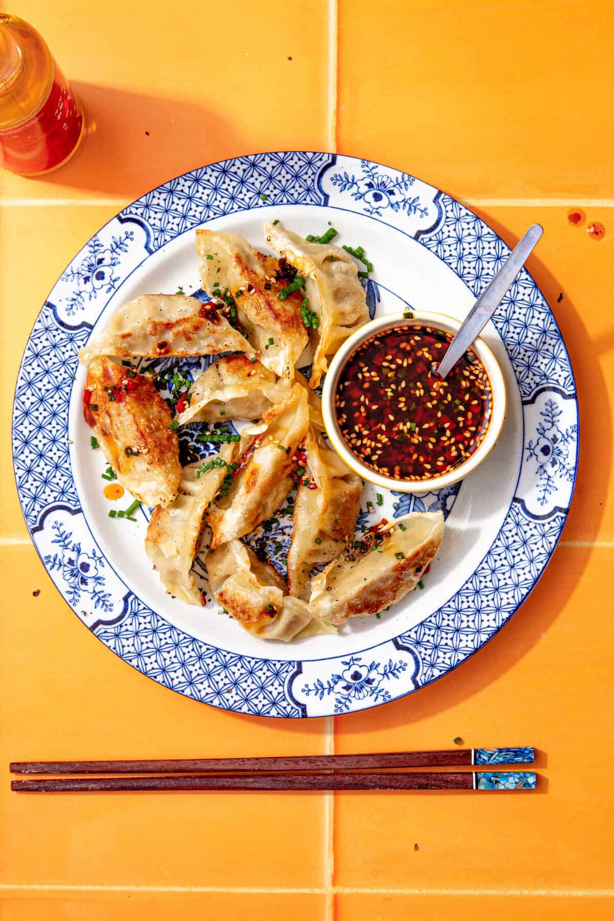 Plate of golden pan-fried gyoza served with a small bowl of easy gyoza dipping sauce on an orange table.